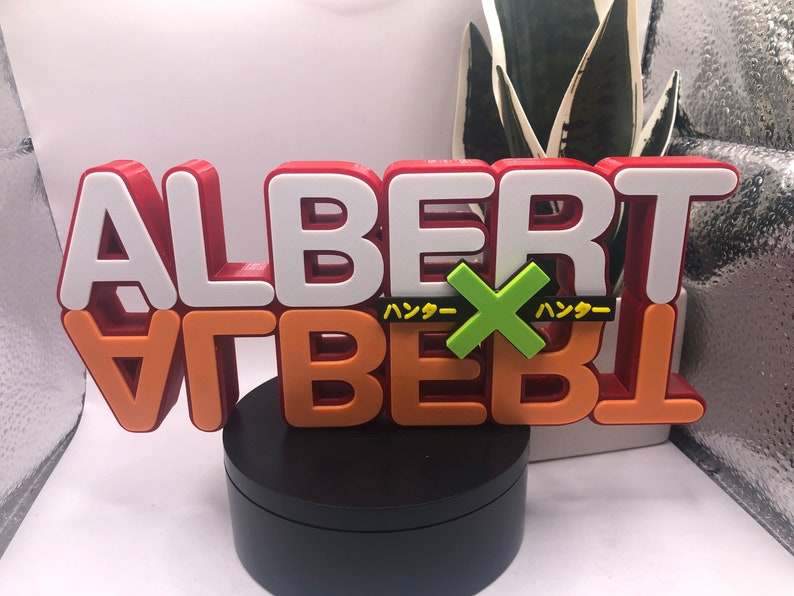 May include: A 3D printed red, white, and orange sign with the word "ALBERT" in white letters on top of the word "ALBERT" in orange letters. The sign has a green "X" with the Japanese word "ハッダー" on either side of the "X". The sign is on a black circular base.
