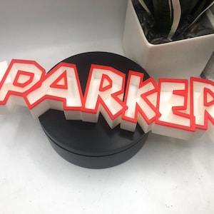 Könnte beinhalten: Ein weißer, 3D-gedruckter Namensschild mit roter Umrandung, auf dem "PARKER" steht.