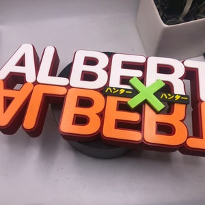 May include: A 3D printed sign with the word "ALBERT" in white letters on top of orange letters with a red outline. The word "ALBERT" is repeated in orange letters with a red outline. A green "X" is in the middle of the sign with the Japanese word "ハッター" on either side of the "X".