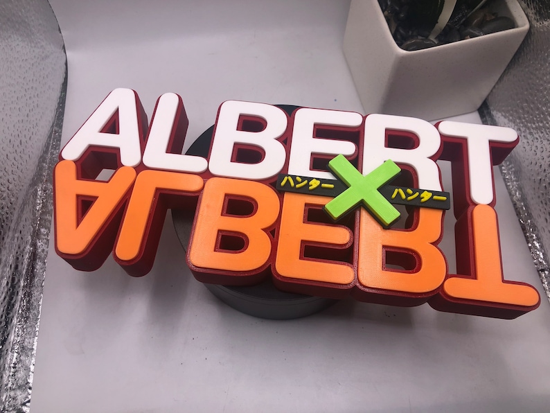 May include: A 3D printed sign with the word "ALBERT" in white letters on top of orange letters with a red outline. The word "ALBERT" is repeated in orange letters with a red outline. A green "X" is in the middle of the sign with the Japanese word "ハッター" on either side of the "X".