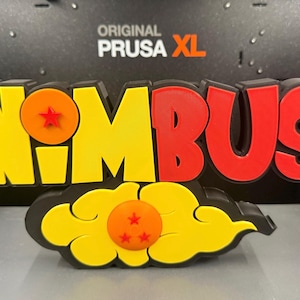 May include: A 3D-printed "Nimbus" sign in yellow and red, with a black outline. The word "Nimbus" is in bold letters, with a star inside the "O". Below the word is a yellow cloud with an orange ball and three stars. The background says "ORIGINAL PRUSA XL".