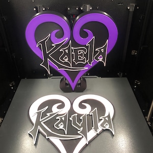 May include: Two heart-shaped nameplates, one purple and black, the other black and white, each with a name in a stylized font. The nameplates are on a gray surface, with a 3D printer in the background.