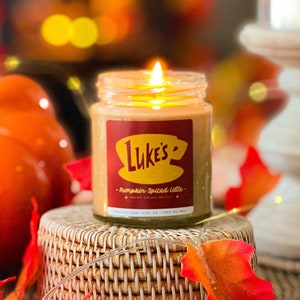 May include: A glass jar candle with a red and yellow label that says "Luke's - Pumpkin Spiced Latte" and a picture of a coffee cup. The candle is sitting on a woven wicker coaster with fall leaves.