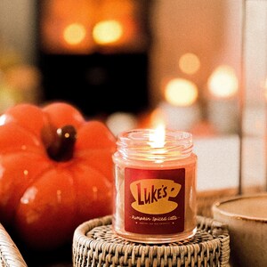 May include: A candle in a glass jar with a red label that says "Luke's" and "Pumpkin Spiced Latte". The candle is lit and sitting on a woven wicker coaster.