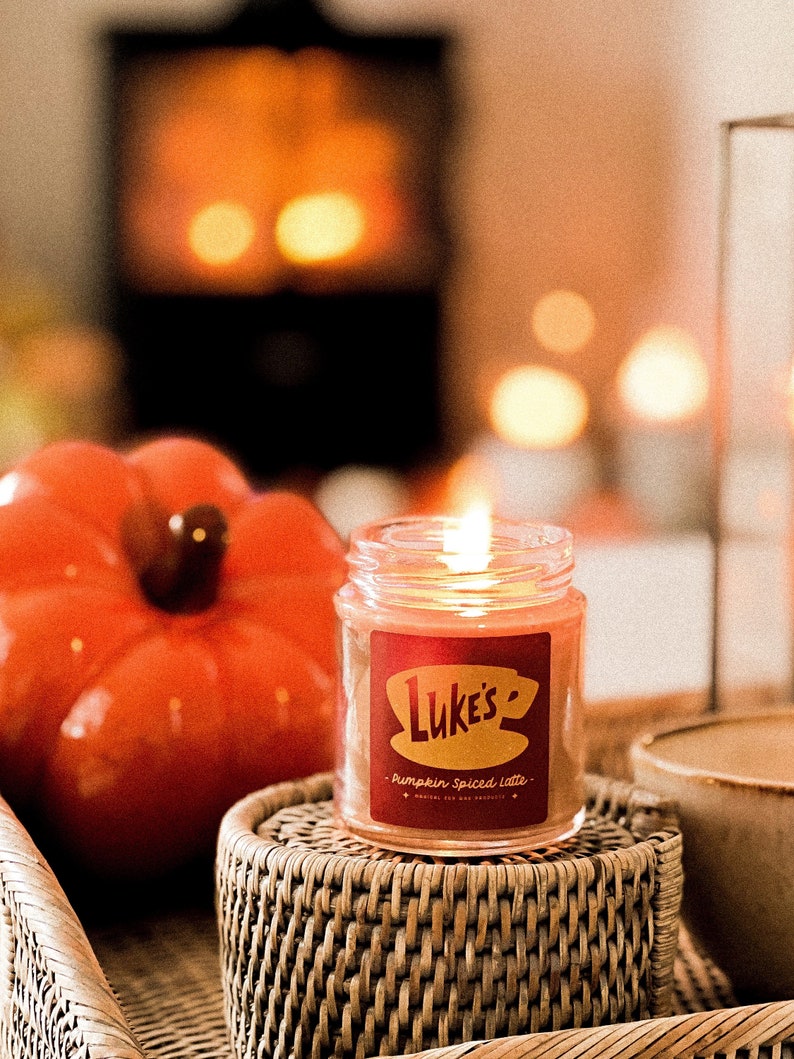 May include: A candle in a glass jar with a red label that says "Luke's" and "Pumpkin Spiced Latte". The candle is lit and sitting on a woven wicker coaster.