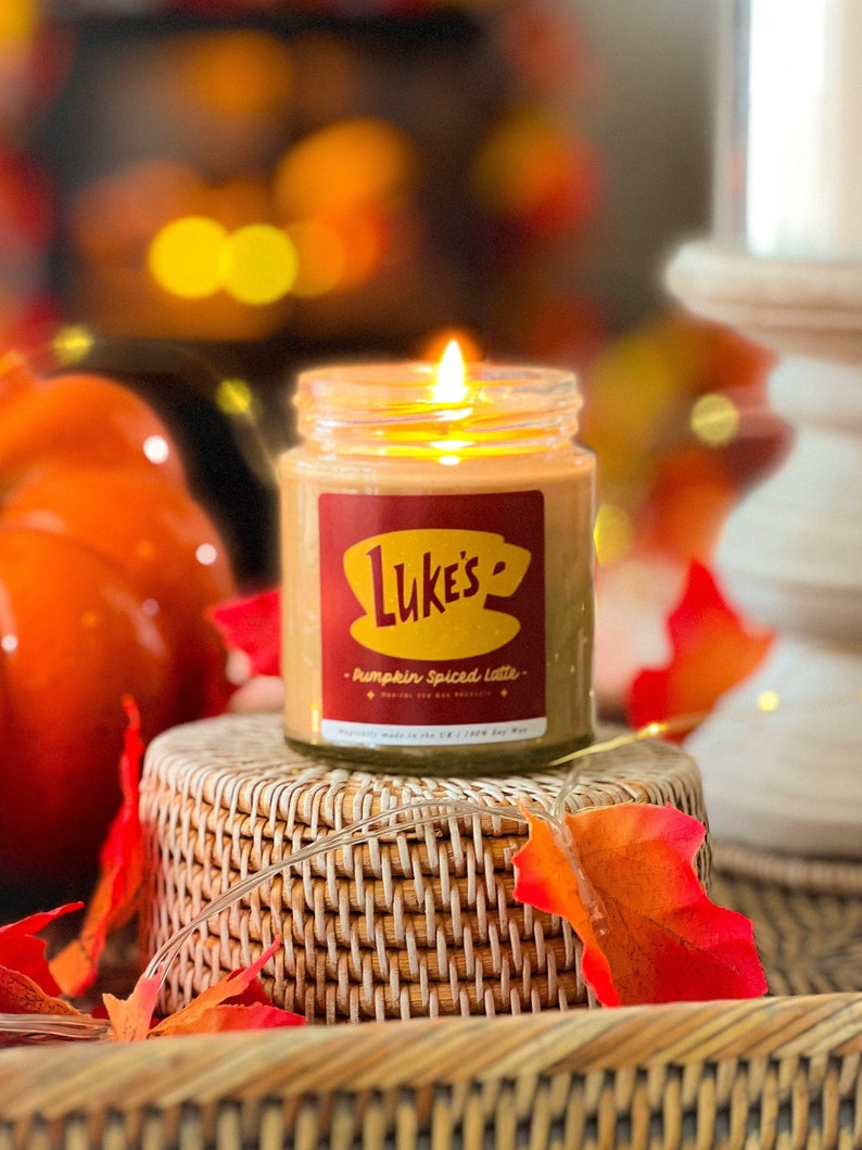 May include: A glass jar candle with a red and yellow label that says "Luke's - Pumpkin Spiced Latte" and a picture of a coffee cup. The candle is sitting on a woven wicker coaster with fall leaves.