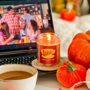 May include: A lit candle with a red label that says "Luke's Pumpkin Spiced Latte" is on a table with a laptop, a mug of coffee, and several orange pumpkins.