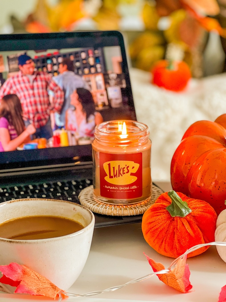 May include: A lit candle with a red label that says "Luke's Pumpkin Spiced Latte" is on a table with a laptop, a mug of coffee, and several orange pumpkins.