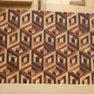 May include: A rectangular wooden chopping board featuring a geometric cube pattern. The design uses various shades of brown wood, creating a 3D effect. The board is likely for kitchen use, with dimensions in centimetres.