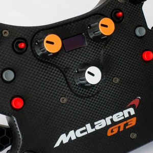 May include: A black racing steering wheel with orange and white accents. The wheel has a carbon fiber finish and features a McLaren GT3 logo. There are several buttons and knobs on the wheel.