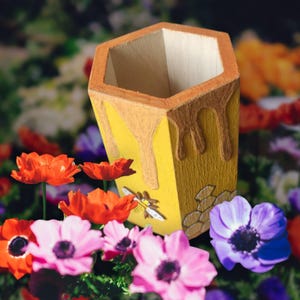 May include: A yellow hexagonal pencil holder with a honey drip design and a bee illustration. The holder is surrounded by colourful flowers.