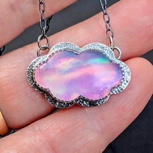 May include: A cloud-shaped pendant necklace with a shimmering pink and purple opal center. The pendant is framed by a silver-toned, textured border and hangs from a delicate chain. The necklace is a unique piece of jewelry.