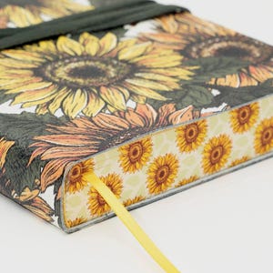 May include: A hardcover journal featuring a sunflower design. The cover displays large yellow and orange sunflowers with green leaves against a white background. A yellow ribbon bookmark and a dark green ribbon closure are visible.