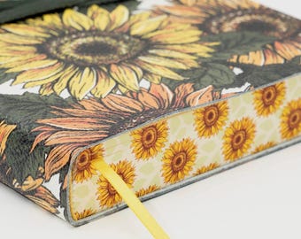 May include: A hardcover journal featuring a sunflower design. The cover displays large yellow and orange sunflowers with green leaves against a white background. A yellow ribbon bookmark and a dark green ribbon closure are visible.