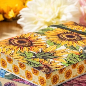 May include: A stack of notebooks with floral designs. The top notebook features a vibrant sunflower pattern with yellow and green hues. The edges are decorated with small sunflower motifs. A black elastic band secures the notebook.