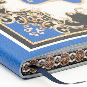 May include: A blue leather-bound journal with a decorative design featuring gold, black, and white baroque elements. The journal has a blue ribbon bookmark and a patterned edge.