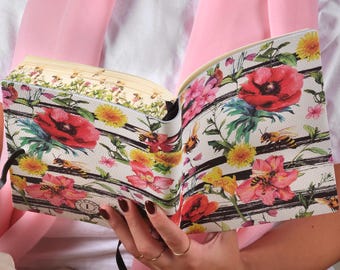 Può includere: Un diario aperto con un design floreale con papaveri rossi, denti di leone gialli e api su sfondo bianco a strisce nere. Il diario ha un nastro nero.