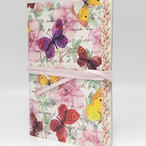 May include: A pink and white journal with a floral design featuring butterflies and handwritten text. The journal is tied with a pink ribbon.