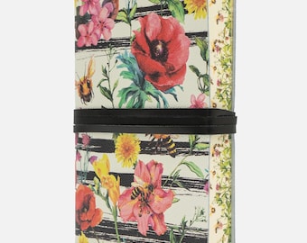 Op de afbeelding: Een bloemenjournaal met een zwarte elastische band. De omslag heeft een crèmekleurige achtergrond met zwarte strepen en kleurrijke bloemen, waaronder rode klaprozen, roze bloesems en gele paardenbloemen. Er zijn ook bijen afgebeeld.