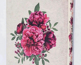 Puede incluir: Un libro de tapa dura con una cubierta de textura beige claro que presenta una ilustración central de peonías de color rosa intenso y hojas verdes. Los bordes de las páginas están decorados con un patrón floral continuo de flores rosas y rojas sobre un fondo crema.