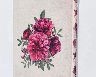 May include: A hardcover book with a textured light beige cover featuring a central illustration of vibrant magenta and deep pink peony flowers with green leaves. The page edges are decorated with a repeating floral pattern of pink and red blossoms.