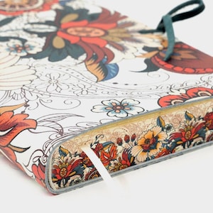 May include: A floral patterned journal with a white background and colorful flowers in red, orange, and blue. The journal has a gray border and a green tie closure. A white ribbon bookmark is visible.