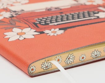 May include: Coral-colored notebook featuring a vintage typewriter illustration with floral accents. The notebook's cover is adorned with white daisies and brown leaves. A white ribbon bookmark is visible, and the edges are decorated with floral patterns.