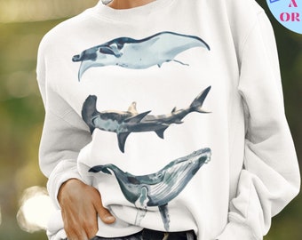 May include: White sweatshirt featuring watercolor illustrations of marine life: a manta ray, a hammerhead shark, and a whale. The sweatshirt has a round neck and long sleeves. Text on the upper right says "FOR AN OVERSIZED FIT + GO UP A SIZE OR TWO!"