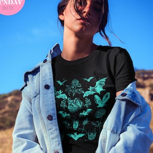 May include: A person wearing a black t-shirt with a teal green graphic print of bats and flowers. The graphic print is reminiscent of vintage botanical illustrations. The person is wearing a blue denim jacket and is standing in front of a blue sky and a brown hillside.