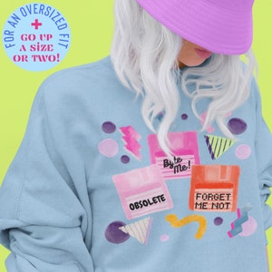 Peut inclure: Un sweat-shirt bleu clair avec un motif rétro représentant des disquettes avec le texte "Byte Me !", "Obsolete" et "Forget Me Not".