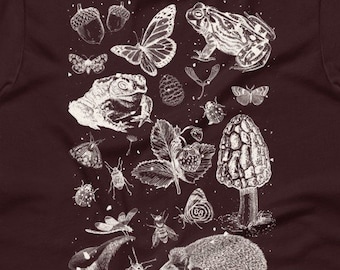 May include: A dark brown t-shirt with a white print of various forest creatures, including a frog, butterfly, mushroom, hedgehog, and insects. The design is a whimsical and detailed illustration.