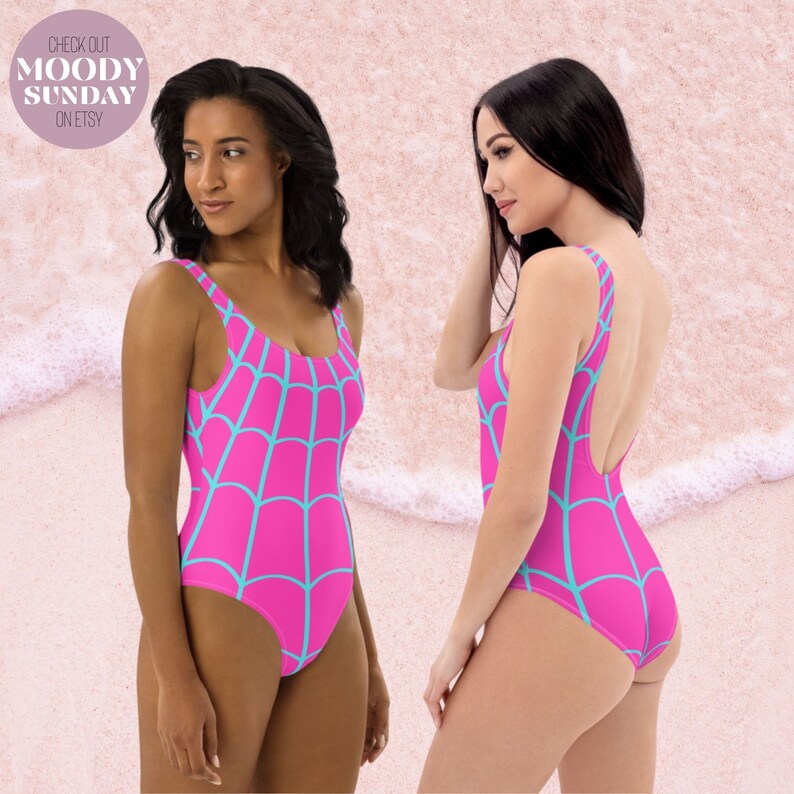May include: A one-piece swimsuit featuring a vibrant hot pink base colour with a repeating light blue spiderweb pattern. The garment has a scoop neckline, wide shoulder straps, and a high-cut leg design, shown from both the front and side angles.