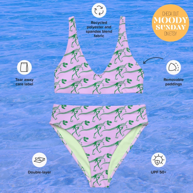 K&ouml;nnte beinhalten: Ein zweiteiliger Bikini in Hellrosa mit einem sich wiederholenden Muster aus gr&uuml;nen Dinosaurierskeletten. Das Oberteil hat einen Rundhalsausschnitt und das Unterteil ist hoch geschnitten. Merkmale sind ein Stoff aus recyceltem Polyester-Elasthan-Gemisch, ein abrei&szlig;bares Pflegeetikett, herausnehmbare Polster, eine doppelte Stofflage und UPF 50+ Schutz.