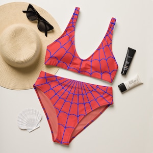 K&ouml;nnte beinhalten: Ein zweiteiliger Bikini aus leuchtend rotem Stoff mit blauem Spinnennetz-Muster. Das Set besteht aus einem Bikini-Oberteil mit Rundhalsausschnitt und einer Bikini-Hose mit hoher Taille. Dargestellt mit einem Strohhut, einer Sonnenbrille und Sonnencreme.