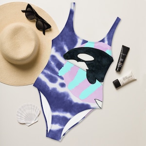 May include: A blue and white tie-dye swimsuit with a black and white orca whale graphic. The orca is swimming in a pink and blue circle.