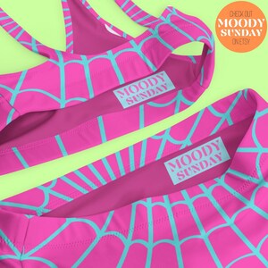 Może przedstawiać: R&oacute;żowy i niebieski str&oacute;j kąpielowy z wzorem pajęczyny. Str&oacute;j kąpielowy ma gorset bandeau i wysokie majtki. Tekst "MOODY SUNDAY" jest nadrukowany na g&oacute;rnej i dolnej części stroju kąpielowego.
