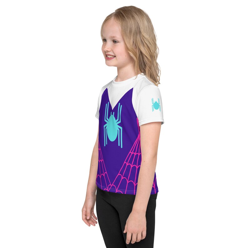 K&ouml;nnte beinhalten: Ein wei&szlig;es T-Shirt mit einem violetten Spinnennetz-Design und einer blauen Spinne in der Mitte. Das Spinnennetz-Design hat rosa Akzente. Das Shirt wird von einem Kind getragen.