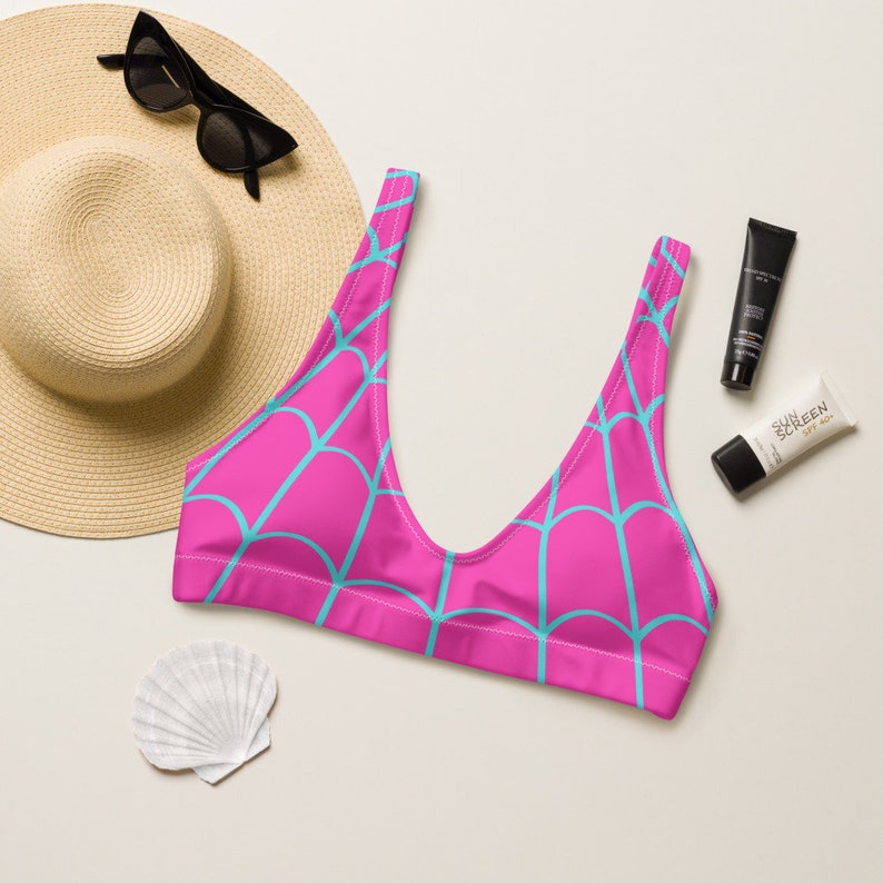 May include: A bright pink bikini top featuring a light blue spiderweb pattern, accompanied by a straw hat, black sunglasses, two tubes of sunscreen, and a white seashell. The bikini top has a deep V-neckline and wide shoulder straps.