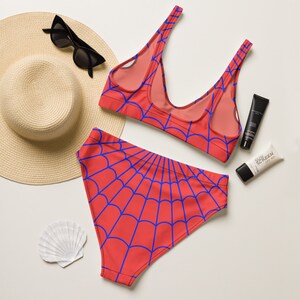 K&ouml;nnte beinhalten: Ein zweiteiliger Bikini mit leuchtend roter Grundfarbe und einem sich wiederholenden blauen Spinnennetz-Muster. Das Set besteht aus einem hoch taillierten Unterteil und einem Oberteil mit Rundhalsausschnitt und breiten Tr&auml;gern, dazu ein Strohhut.