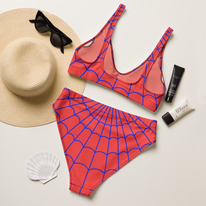 K&ouml;nnte beinhalten: Ein zweiteiliger Bikini mit leuchtend roter Grundfarbe und einem sich wiederholenden blauen Spinnennetz-Muster. Das Set besteht aus einem hoch taillierten Unterteil und einem Oberteil mit Rundhalsausschnitt und breiten Tr&auml;gern, dazu ein Strohhut.