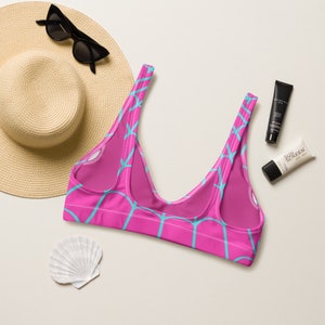 May include: A bright pink bikini top featuring a repeating light blue geometric grid pattern. The garment has wide shoulder straps, a deep scoop neckline, and visible white stitching along the edges of the fabric construction.
