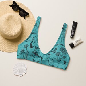May include: A turquoise bikini top with a black and white print of insects, flowers, and a skeleton. The print is a whimsical and slightly macabre design.