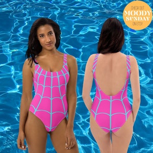 May include: A one-piece swimsuit featuring a vibrant hot pink base colour with a light blue spiderweb pattern across the front and back. The garment has a scoop neckline and wide shoulder straps. Text in the upper right corner reads CHECK OUT MOODY SUNDAY ON ETSY.