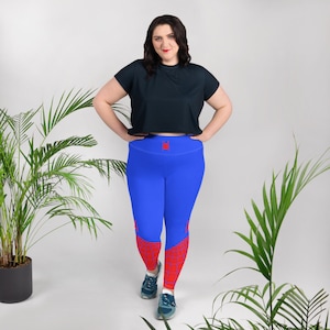 Puede incluir: Una mujer lleva pantalones de yoga azules con un diseño de telaraña roja en la parte inferior de las piernas. Los pantalones de yoga tienen cintura alta y un pequeño logotipo de araña roja en la cintura. Lleva una camiseta negra de manga corta.