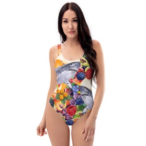 Puede incluir: Traje de baño de una pieza con un diseño de acuarela de un tiburón rodeado de varias frutas, incluyendo naranjas, arándanos y frambuesas. El traje de baño tiene un escote redondo y una pernera de corte alto. Los colores son vibrantes y el diseño es llamativo.