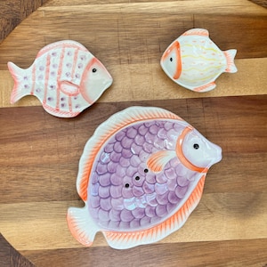 Könnte beinhalten: Drei Keramik-Fischteller. Einer ist lila mit orangefarbenem Rand, einer ist weiß mit orangefarbenen und gelben Streifen und einer ist weiß mit rosa und orangefarbenen Streifen.