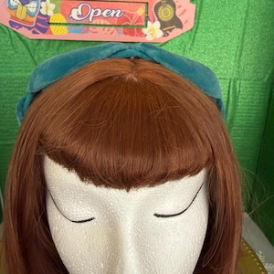 May include: A teal velvet headband sits atop a mannequin head with auburn hair and bangs. The mannequin has closed eyes and red lipstick. A sign in the background reads "Tiki Bar Open".