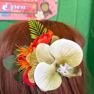 May include: A colorful hair accessory featuring a large, cream-colored orchid flower with green and purple accents. The arrangement includes orange, red, and green artificial flowers and foliage. The hairpiece is set against brown hair.