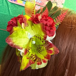May include: A hair accessory featuring a large, vibrant green and red orchid, deep red fabric flowers, and green fern-like foliage. The orchid petals have a textured, speckled appearance with glittery edges, arranged in a cluster with other floral elements.
