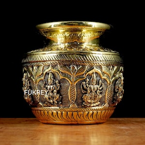 May include: A gold-colored, ornate brass pot with intricate carvings of deities and floral patterns. The pot has a wide base, a rounded body, and a flared rim. The text "Fukrey" is visible on the side.